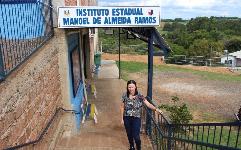 Professora Denise, na entrada da escola, com a placa do antigo prédio do instituto estadual