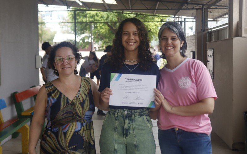 Alice, ao centro, exibe o certificado recebido na Assembleia Legislativa ao lado da diretora da Escola, Andréia Luciana Pereira (direita) e da professora de matemática, Heidi Daiana Machado (esquerda) | abc+
